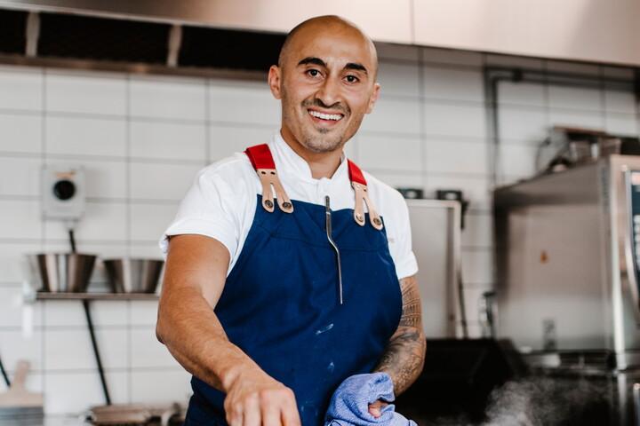Man i förkläde och vit t-short lagar mat i ett restaurangkök. Han tittar in i kameran och ler.